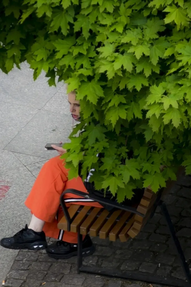 Person in orange trousers seated on a bench hidden by green leaves